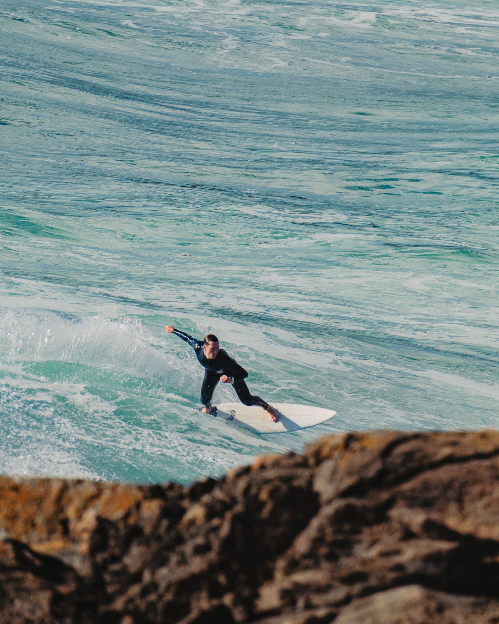Surfeur sur une vague turquoise vu depuis les rochers