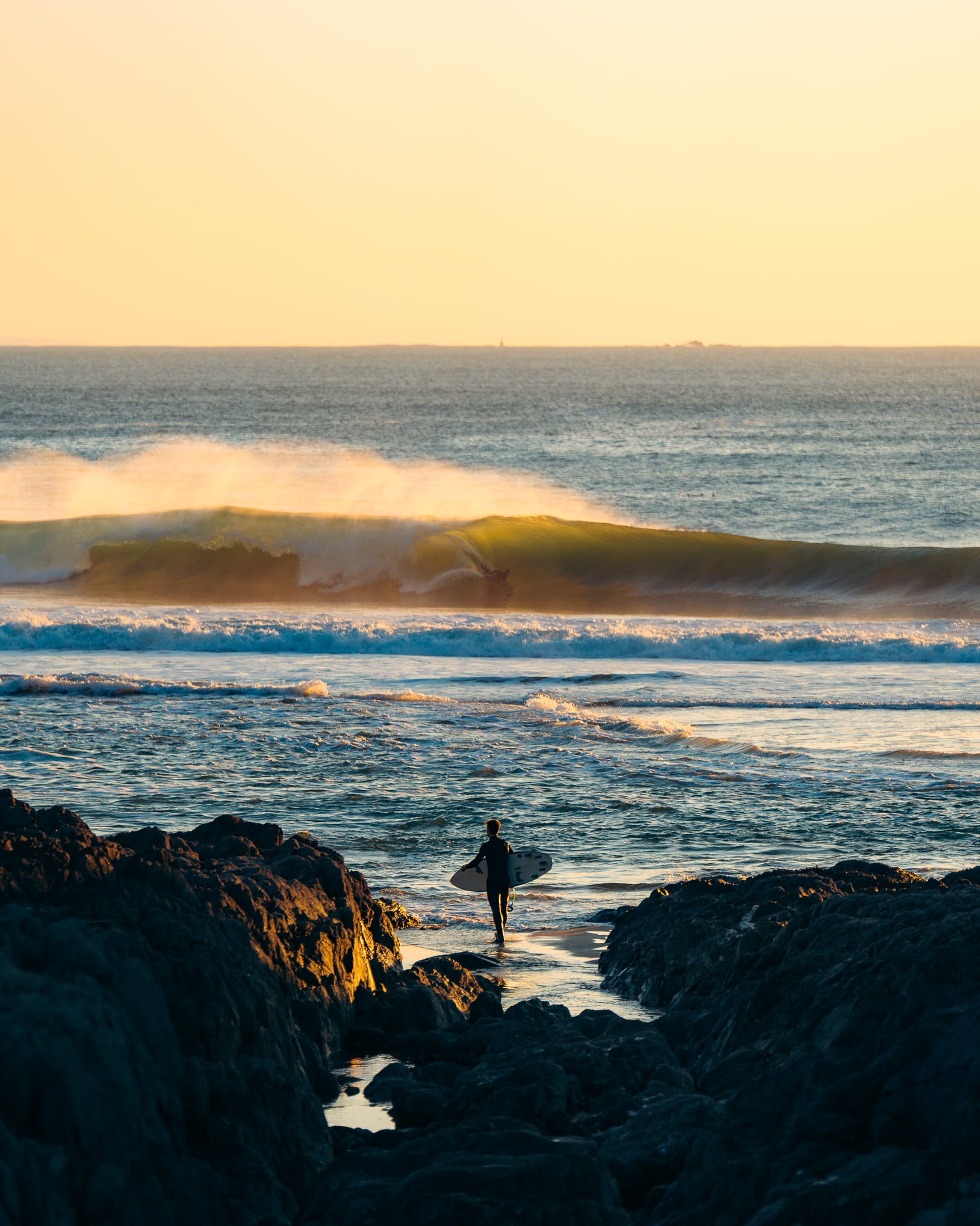 Surfeur marchant sur les rochers au coucher du soleil face aux vagues